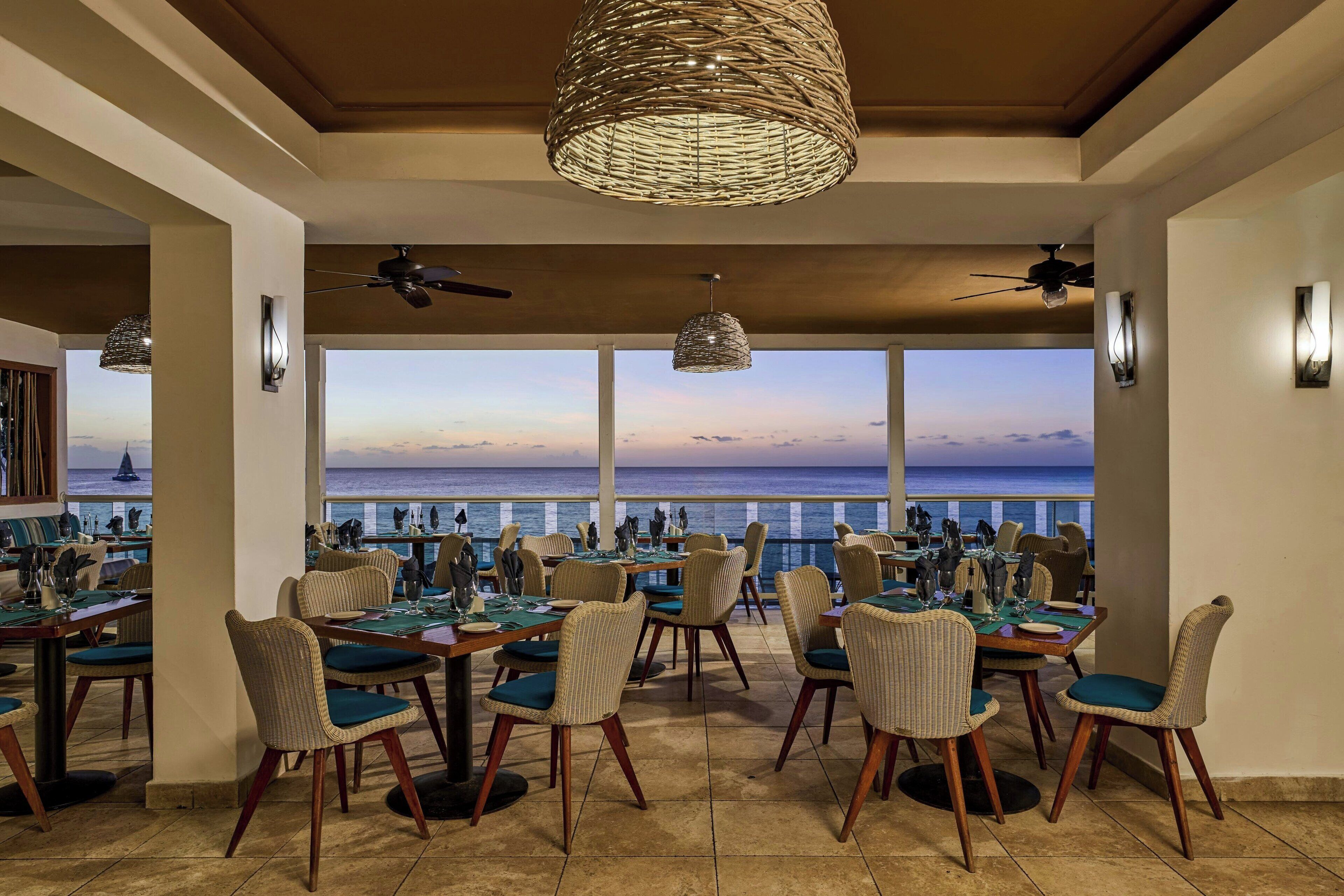 Un restaurante con sillas y mesas de mimbre, amplios ventanales con vistas al mar y ventiladores de techo.