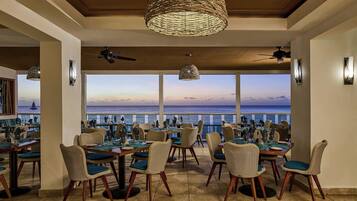 Un restaurante con sillas y mesas de mimbre, amplios ventanales con vistas al mar y ventiladores de techo.