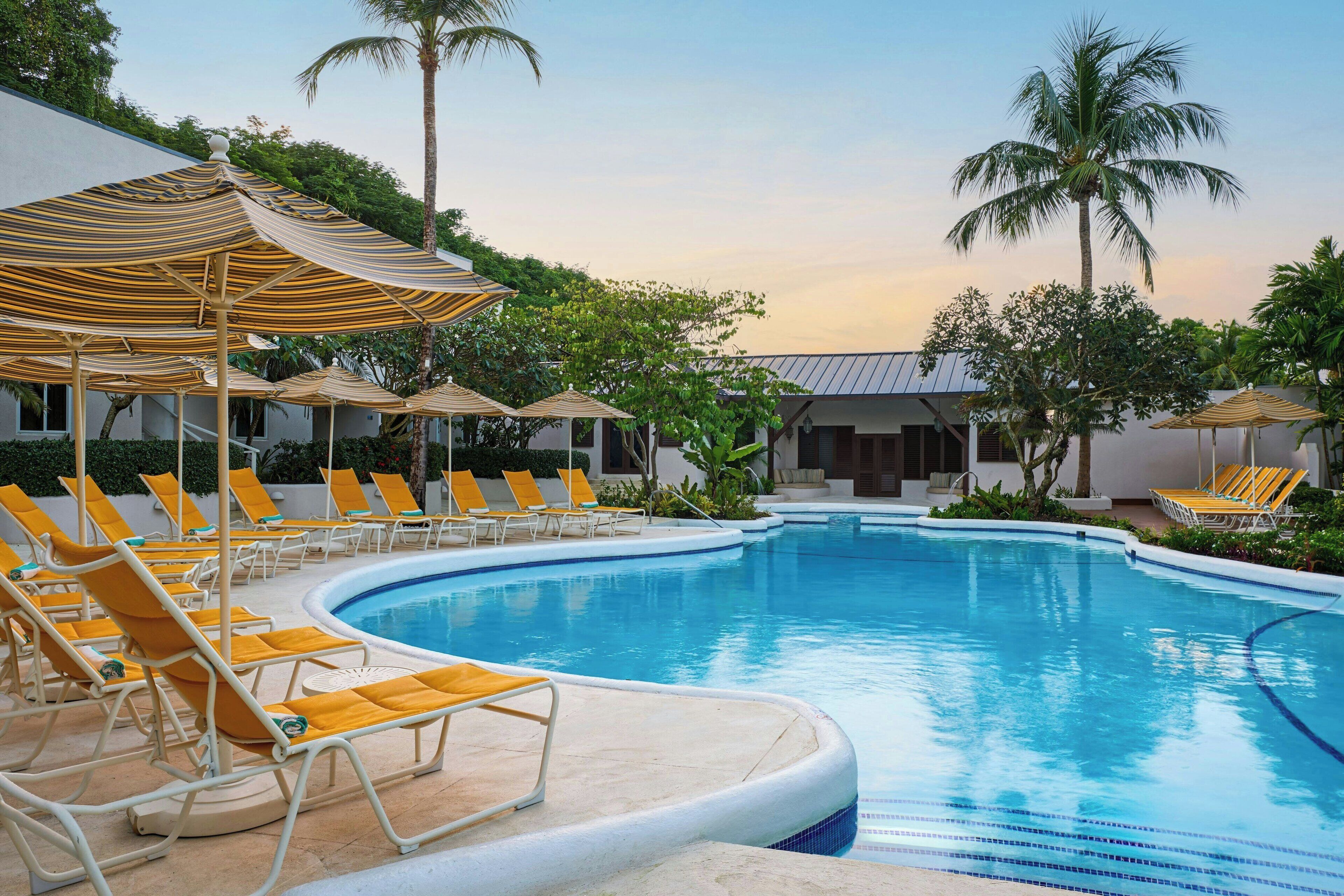 A swimming pool with sun loungers and umbrellas, surrounded by palm trees and a building.