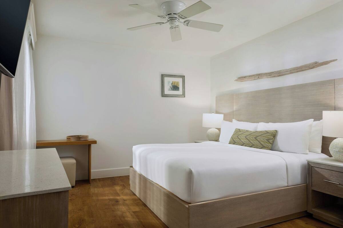 Habitación de hotel ordenada con cama, mesitas de noche, escritorio, ventilador de techo, un cuadro enmarcado en la pared y cabecera de madera.