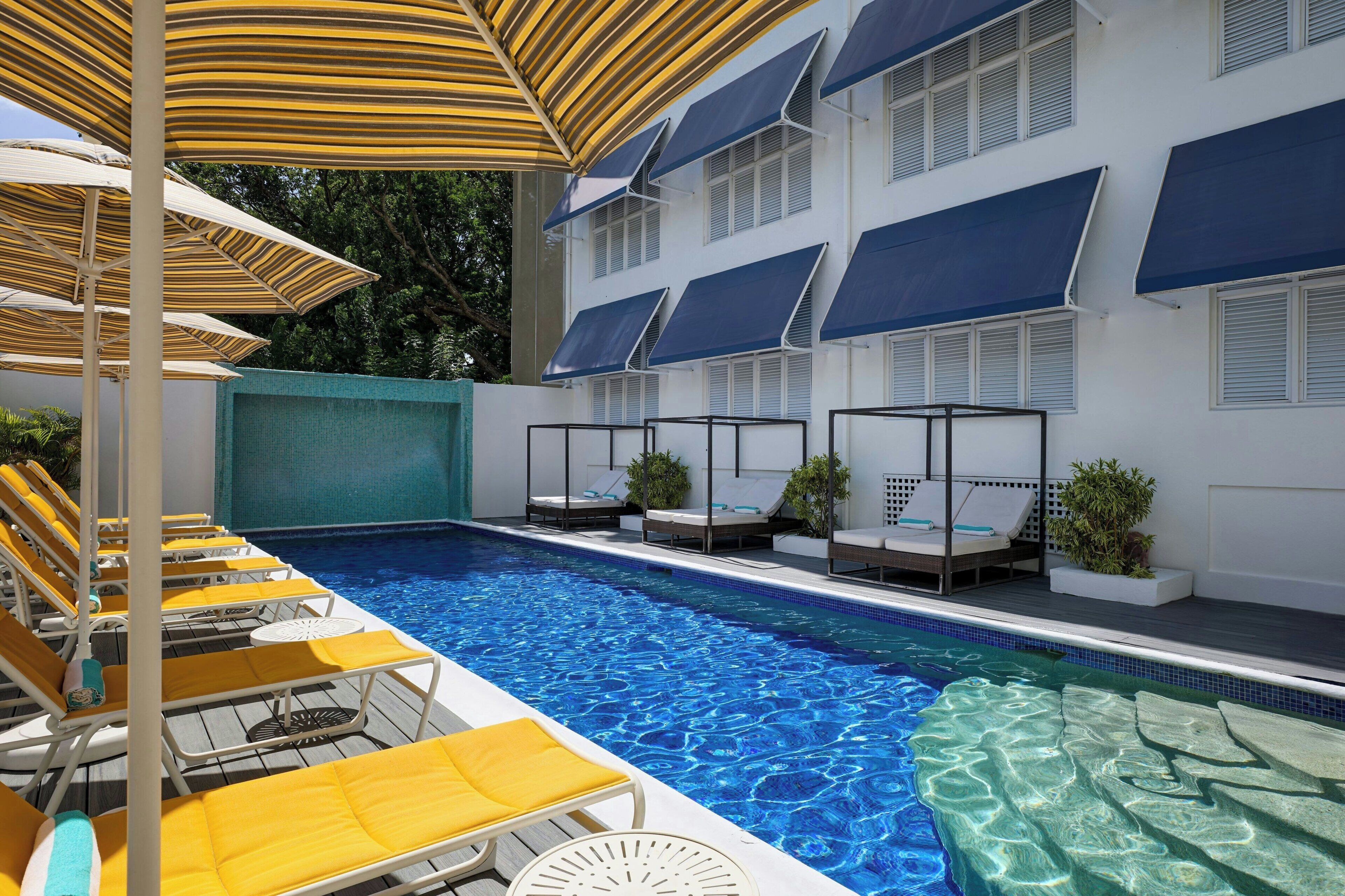 A swimming pool with sun loungers and umbrellas, surrounded by a building with multiple windows and awnings.