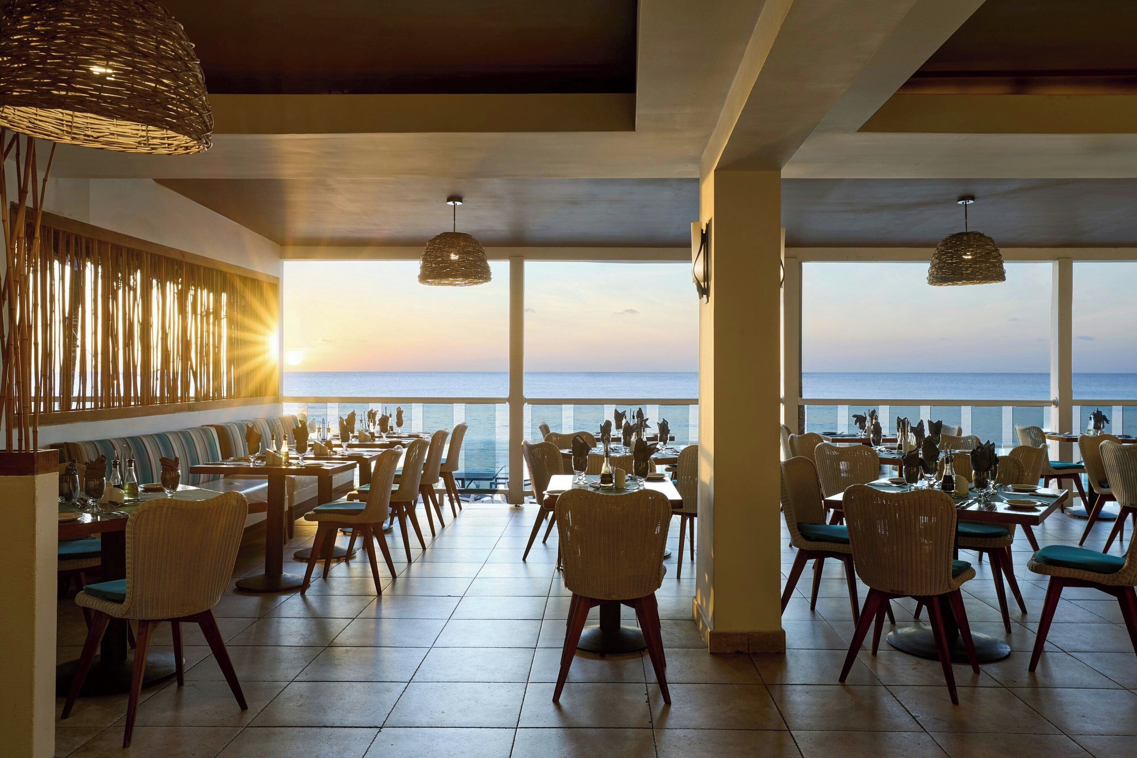 A restaurant with a view of the ocean at sunset, featuring wicker chairs and tables.