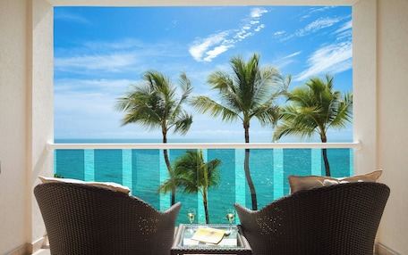 A balcony with wicker chairs, a glass table, and a view of the ocean and palm trees.