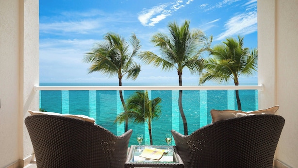 A balcony with wicker chairs, a glass table, and a view of the ocean and palm trees. - Waves Resort & Spa, Barbados, An Autograph Collection® All-Inclusive Resort (Prospect)