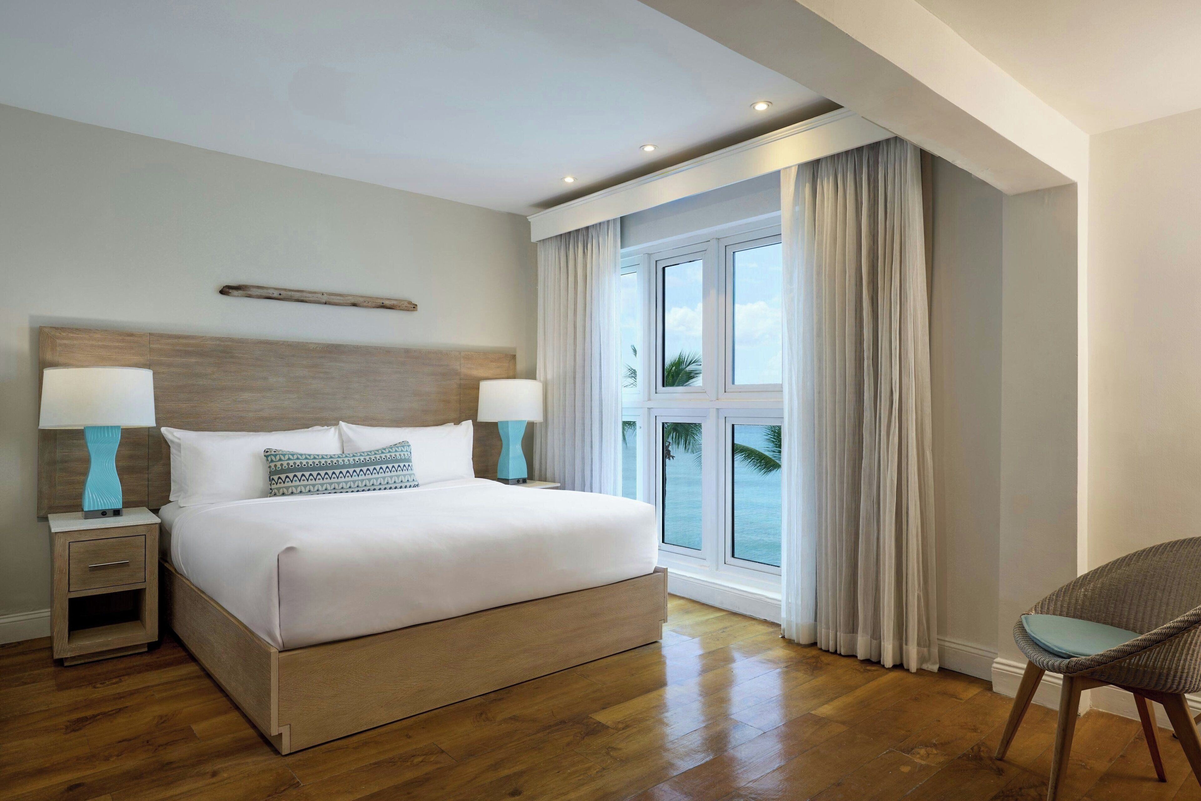 A hotel room with a large bed, two bedside tables with lamps, a view of the ocean through a window, and a chair by the window.