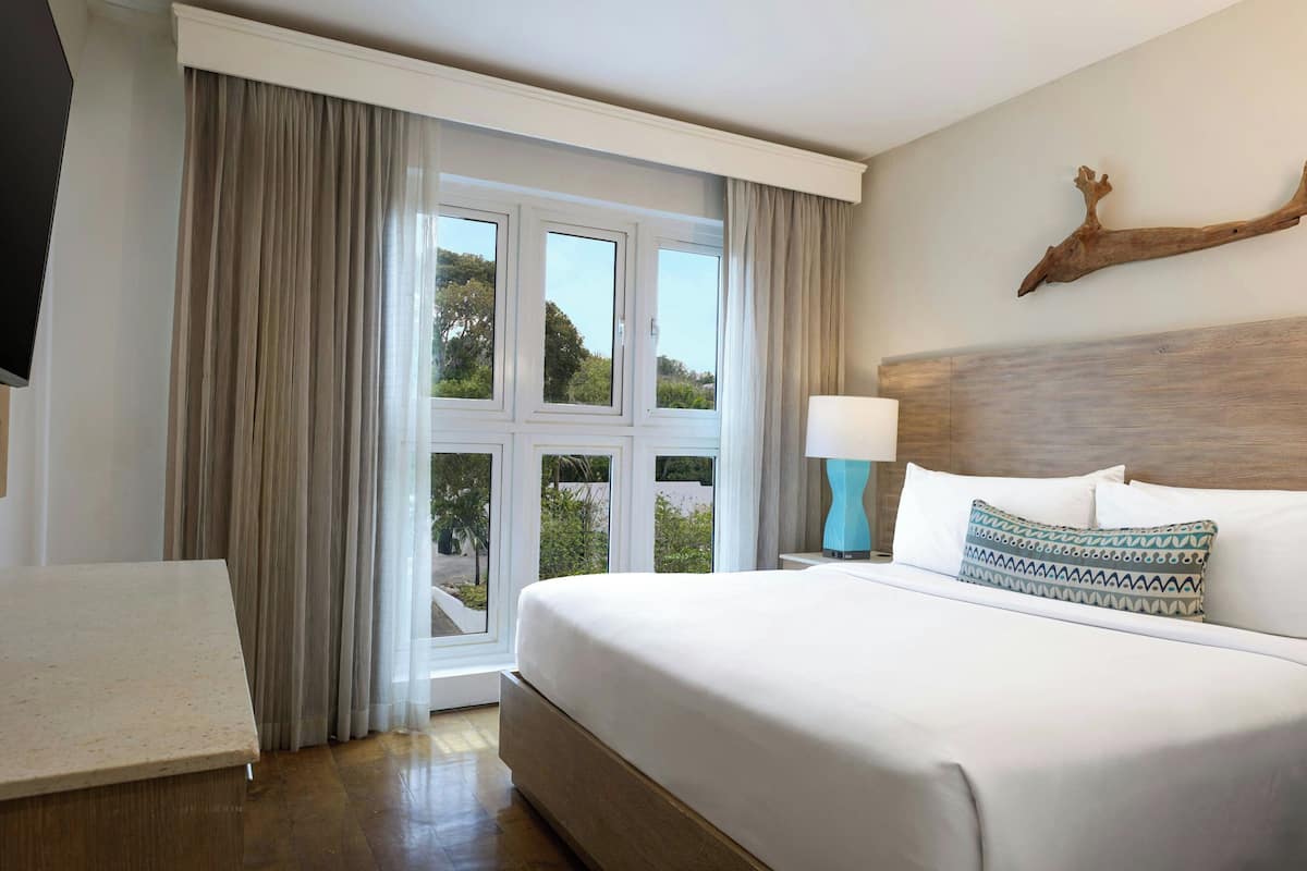 Habitación de hotel con una cama grande, una mesita de noche con lámpara, cabecero de madera y vistas al verdor a través de la ventana.