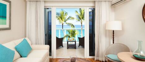 A hotel room with a view of palm trees and the ocean, a white sofa with teal cushions, a wooden coffee table, a wicker chair, and a lamp.