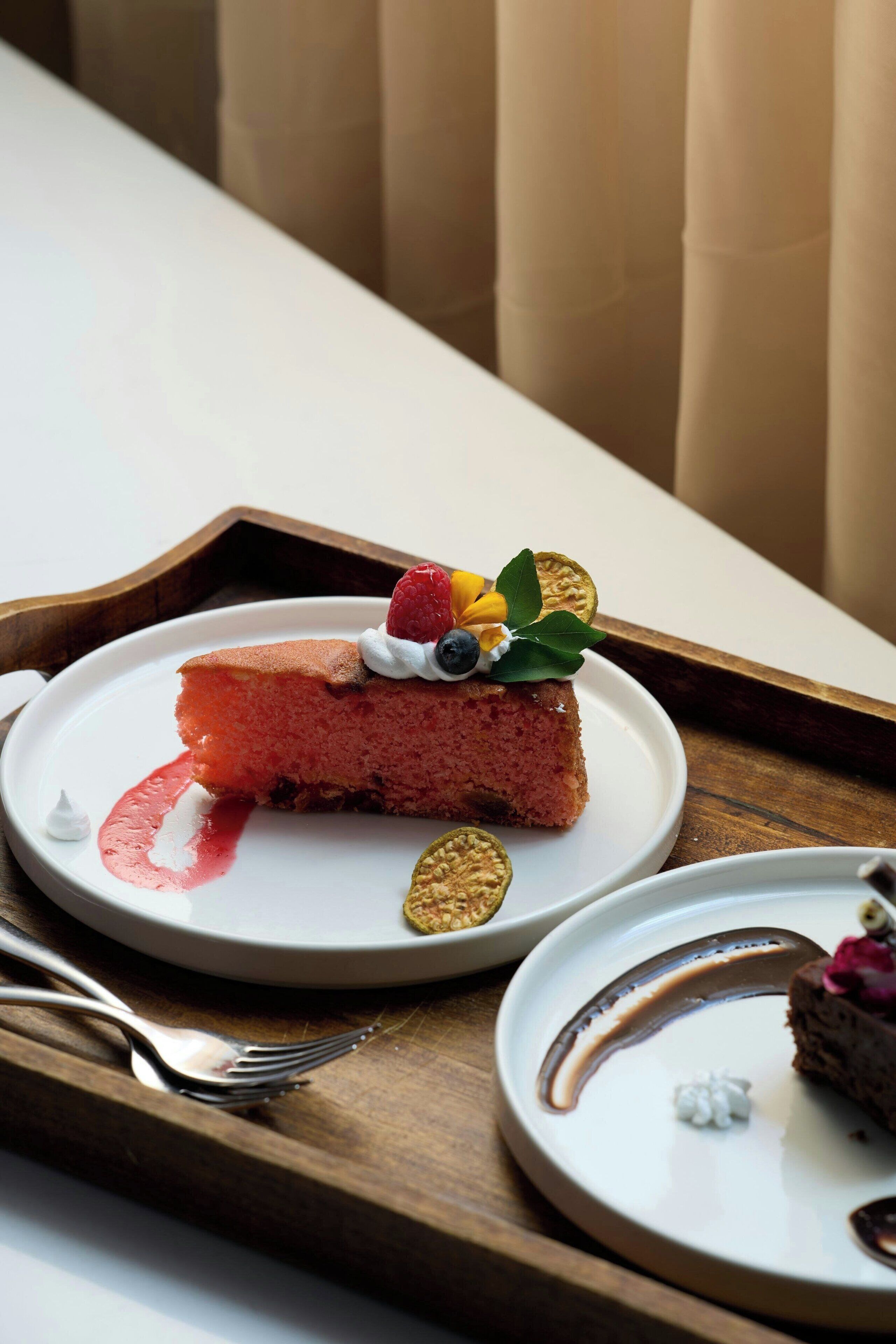 Una porción de pastel de queso en un plato blanco con cobertura de frutos rojos, acompañada de un tenedor y un recipiente pequeño de crema batida.