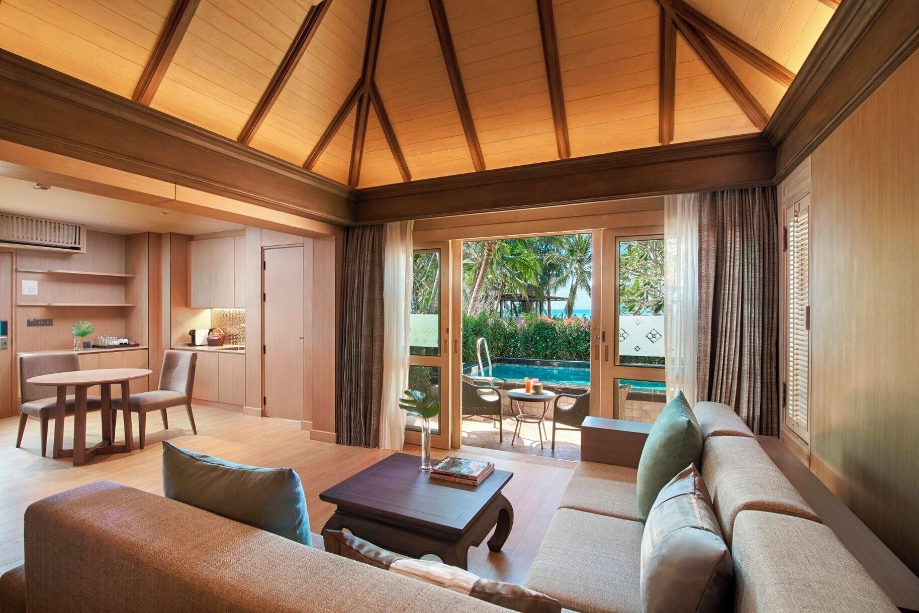 A spacious living area with a large sofa, a coffee table, and a dining table. There is a view of a pool and palm trees through large windows.