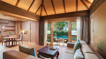 A spacious living area with a large sofa, a coffee table, and a dining table. There is a view of a pool and palm trees through large windows.