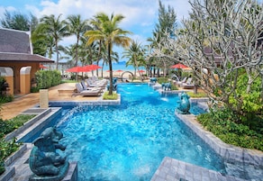 A swimming pool with lounge chairs and umbrellas, surrounded by palm trees and a view of the ocean.