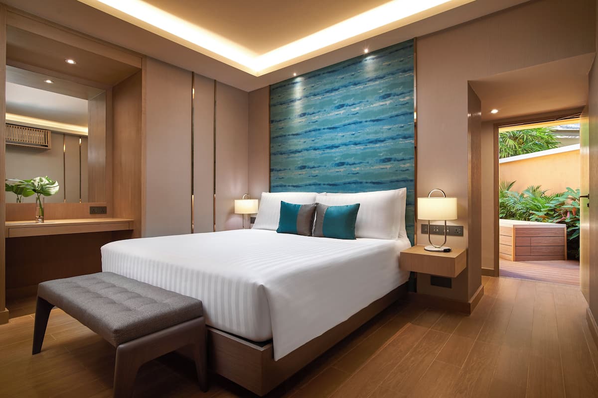 Een moderne hotelkamer met een groot bed, nachtkastjes, een bankje en uitzicht op groen.