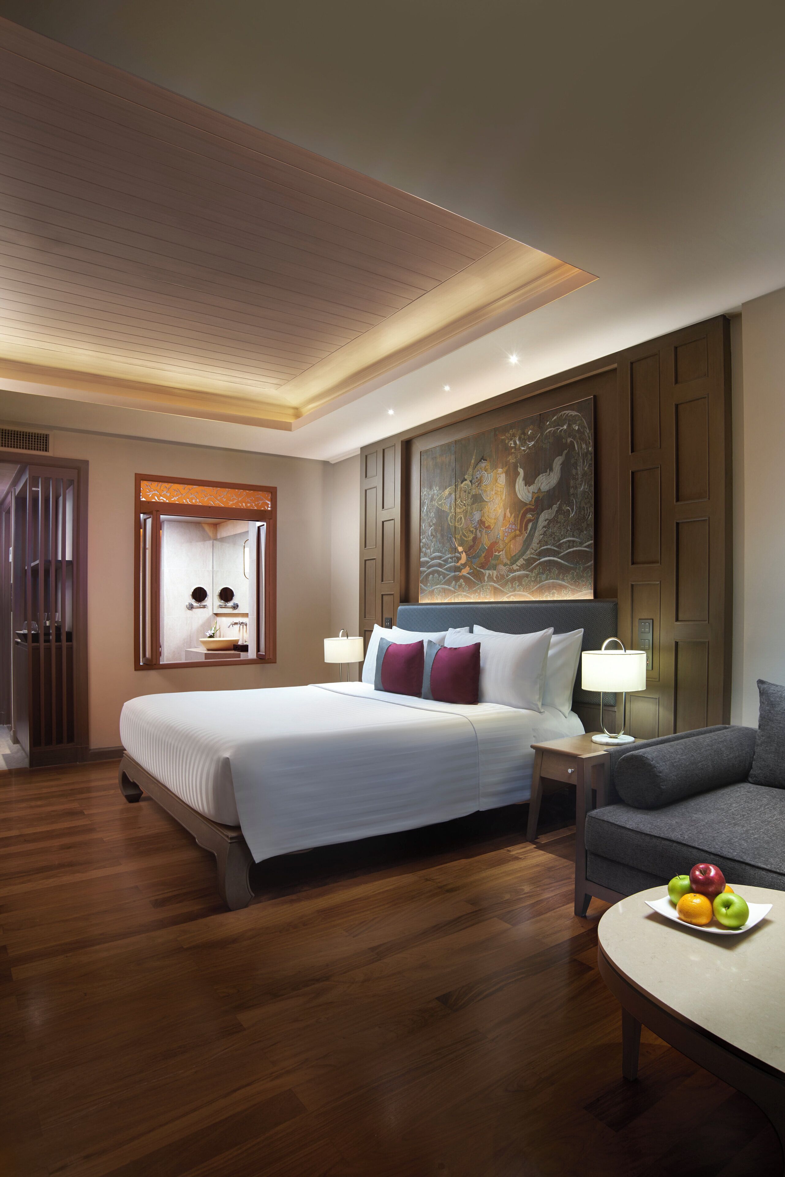 Una habitación de hotel con una cama grande, una mesita de noche con lámpara, un pequeño sofá y una bandeja de frutas.