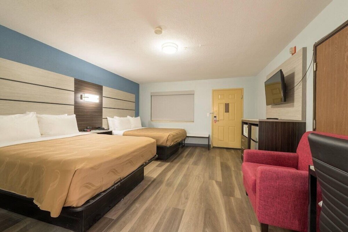 A hotel room with two beds, a TV, a red chair, and a wooden floor.