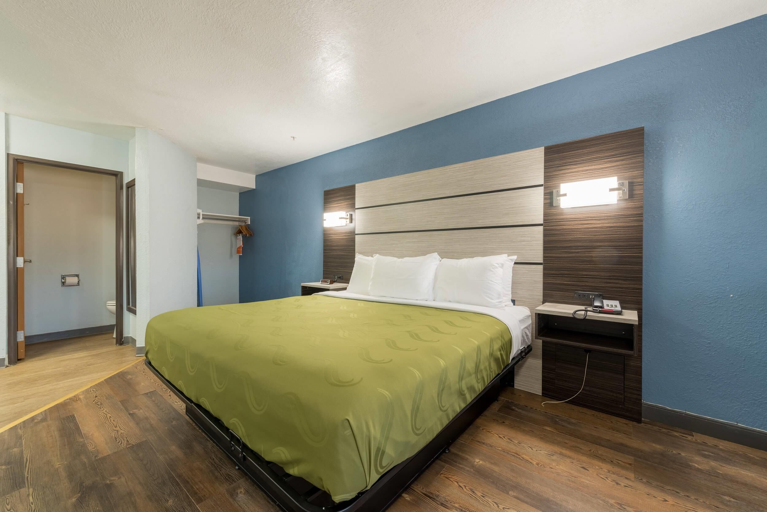 A hotel room with a bed, a nightstand, a wall-mounted light, and a wooden floor.