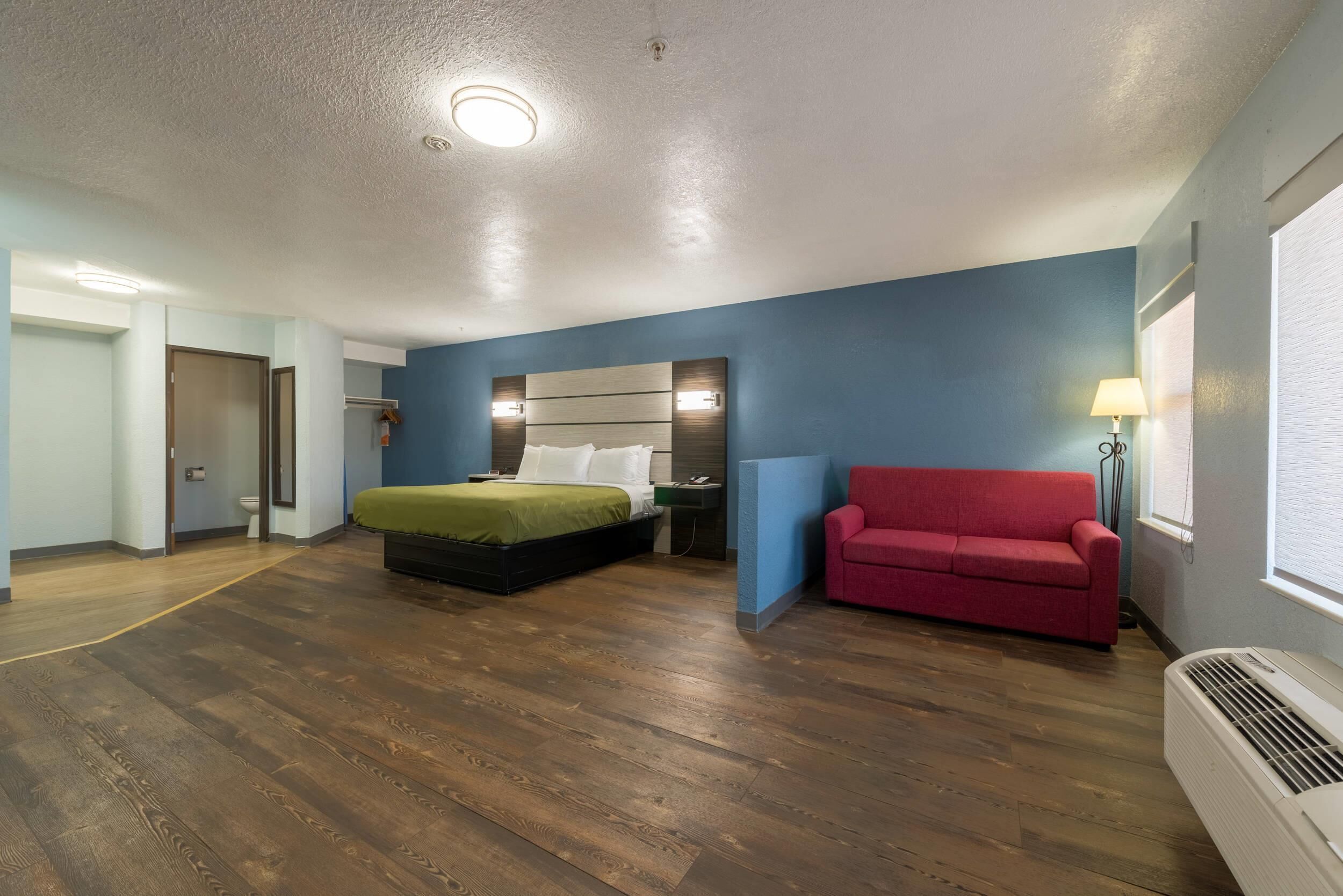 Quarto de hotel com uma cama, um sofá vermelho, uma mesa de cabeceira, um abajur e um ar condicionado.