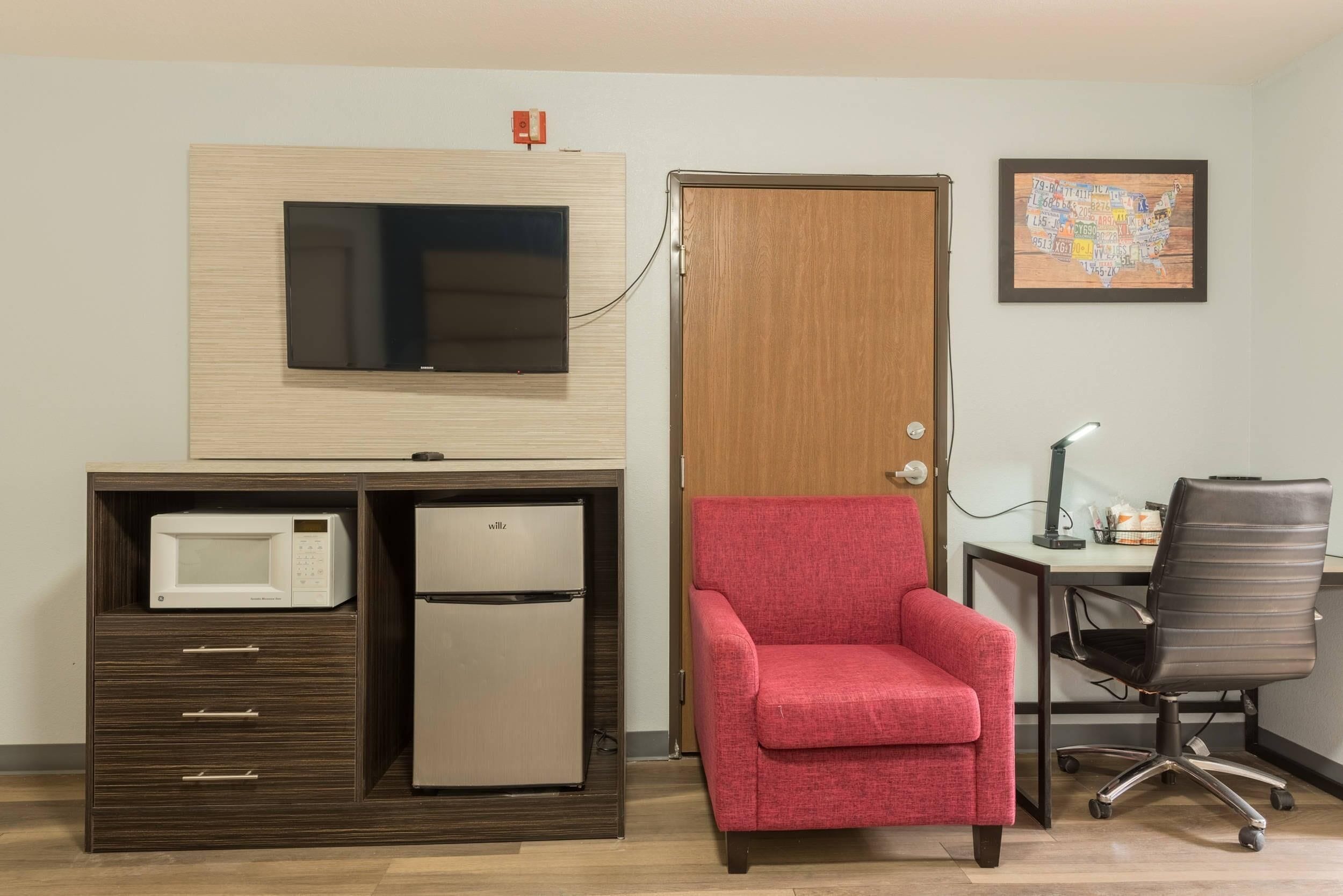 A compact room with a kitchenette, a red armchair, a desk with a computer, and a wall-mounted TV.