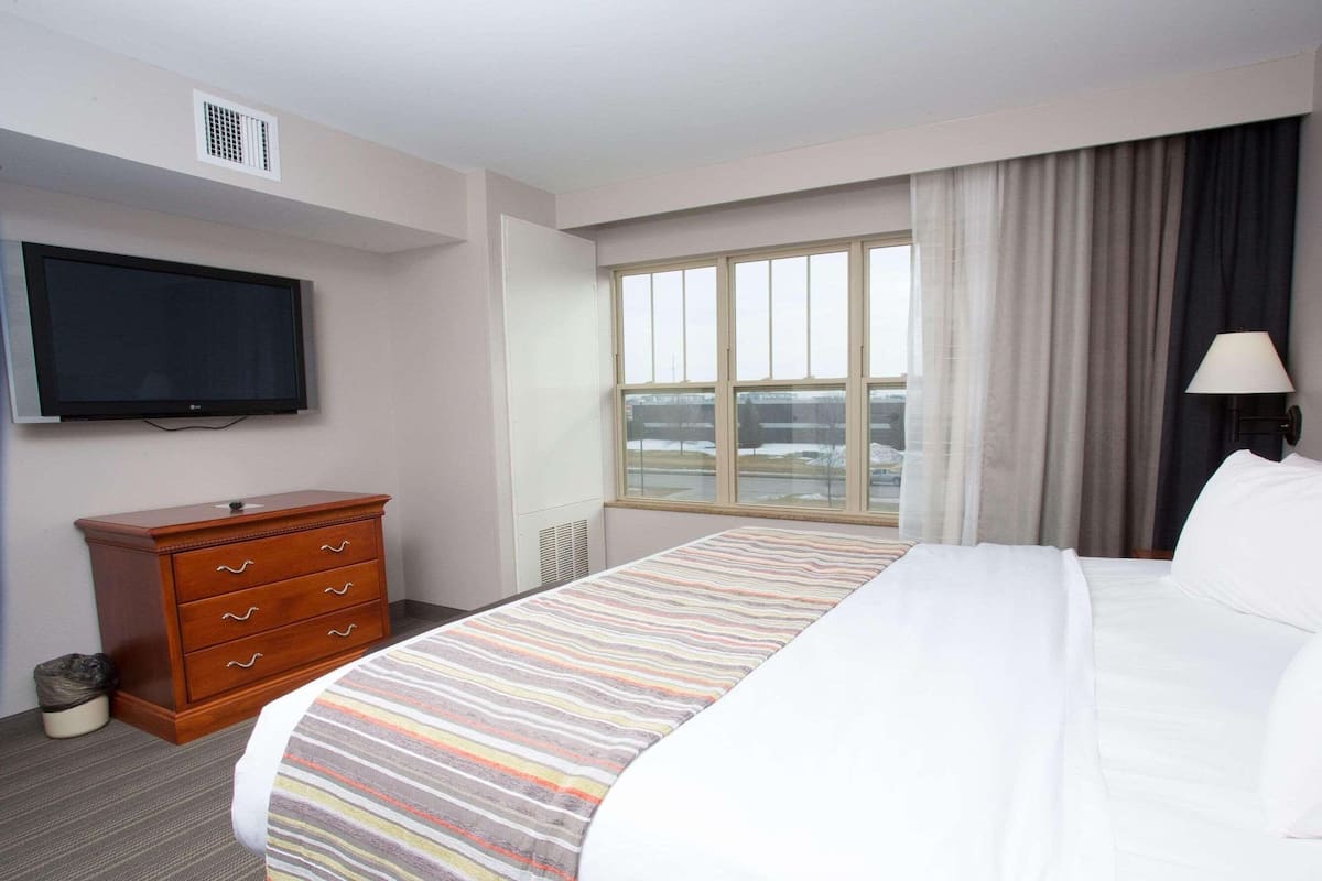 A hotel room with a bed, a dresser, a television, a window with curtains, and a lamp.