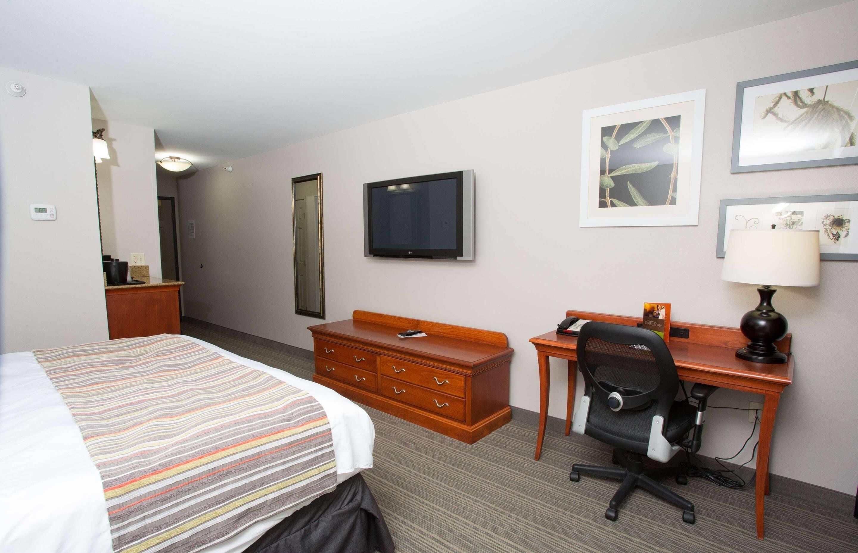 A hotel room with a bed, a wooden dresser, a desk with a chair, a flat-screen TV, and framed artwork on the wall.