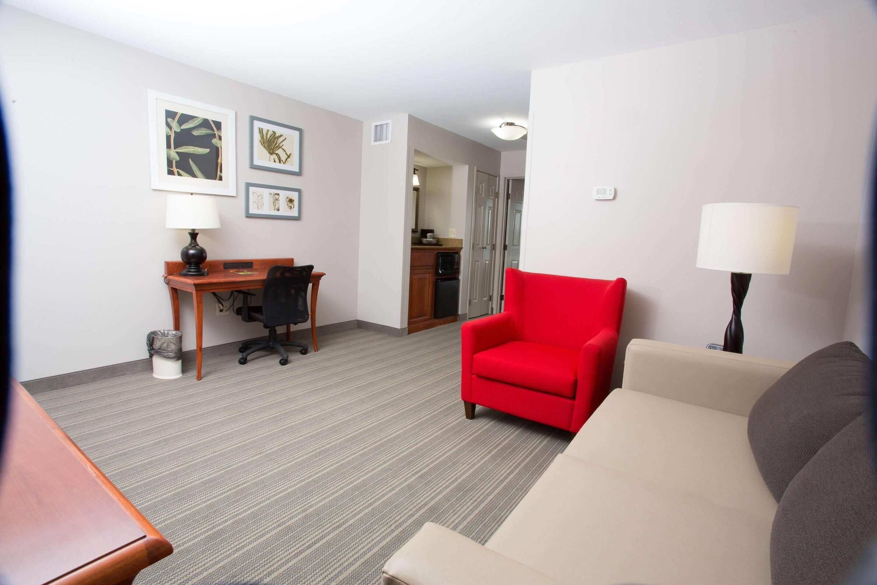 Une chambre d’hôtel avec un fauteuil rouge, un canapé beige, un bureau avec une chaise et des reproductions encadrées de motifs botaniques accrochées au mur.