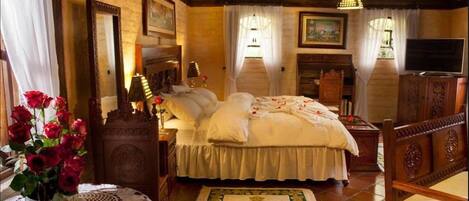 A traditional bedroom with a large bed, wooden headboard, a bedside table with a lamp, a mirror, and a television.