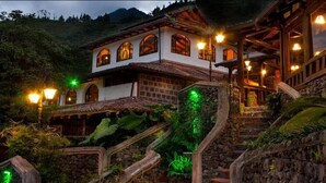 A traditional chalet-style building with a stone staircase leading to a wooden deck, surrounded by lush greenery.