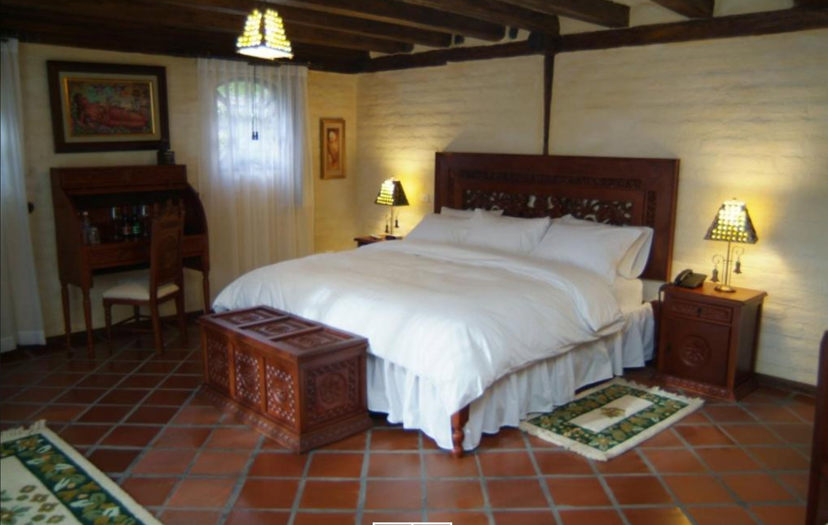 Un dormitorio con una cama grande, cabecera de madera, dos mesitas de noche con lámparas, un banco de madera y suelo de baldosas.
