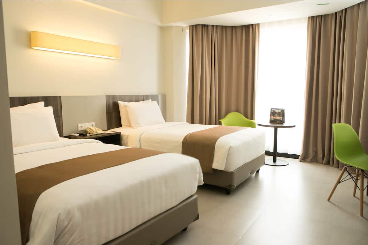 A hotel room with two beds, a small table, a green chair, and a view of the outdoors.