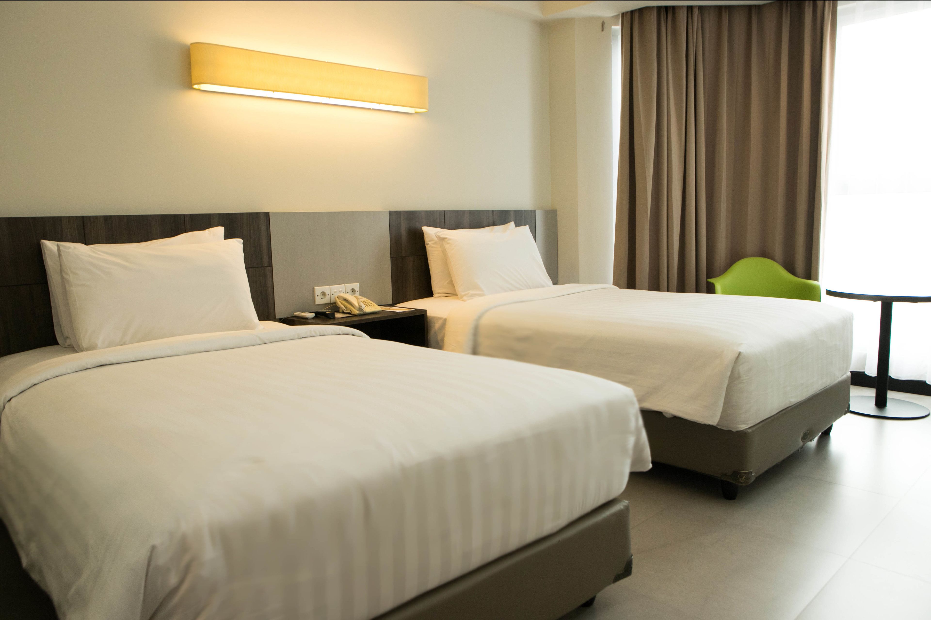 Habitación de hotel con dos camas, una mesita de noche, una lámpara, una silla verde y una ventana con cortinas.