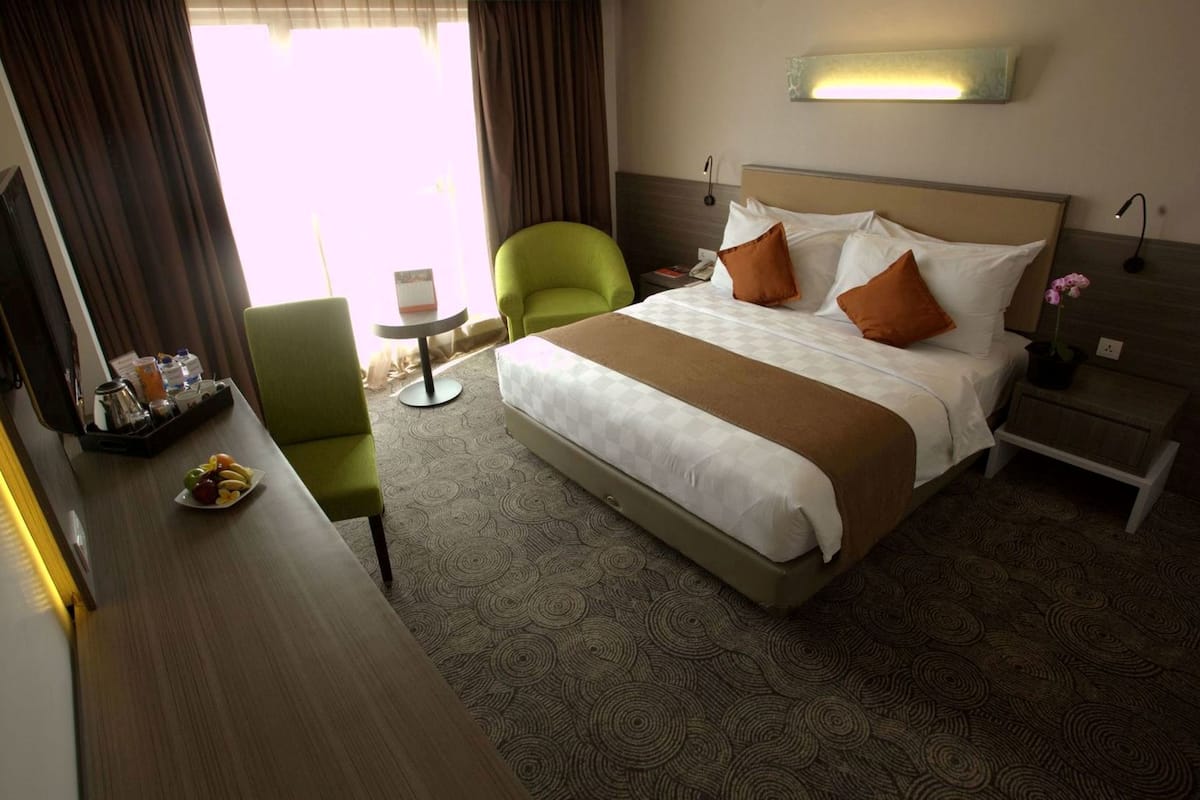 A hotel room with a large bed, a desk with a fruit bowl, a green armchair, a small table with a lamp, and a nightstand with a plant.