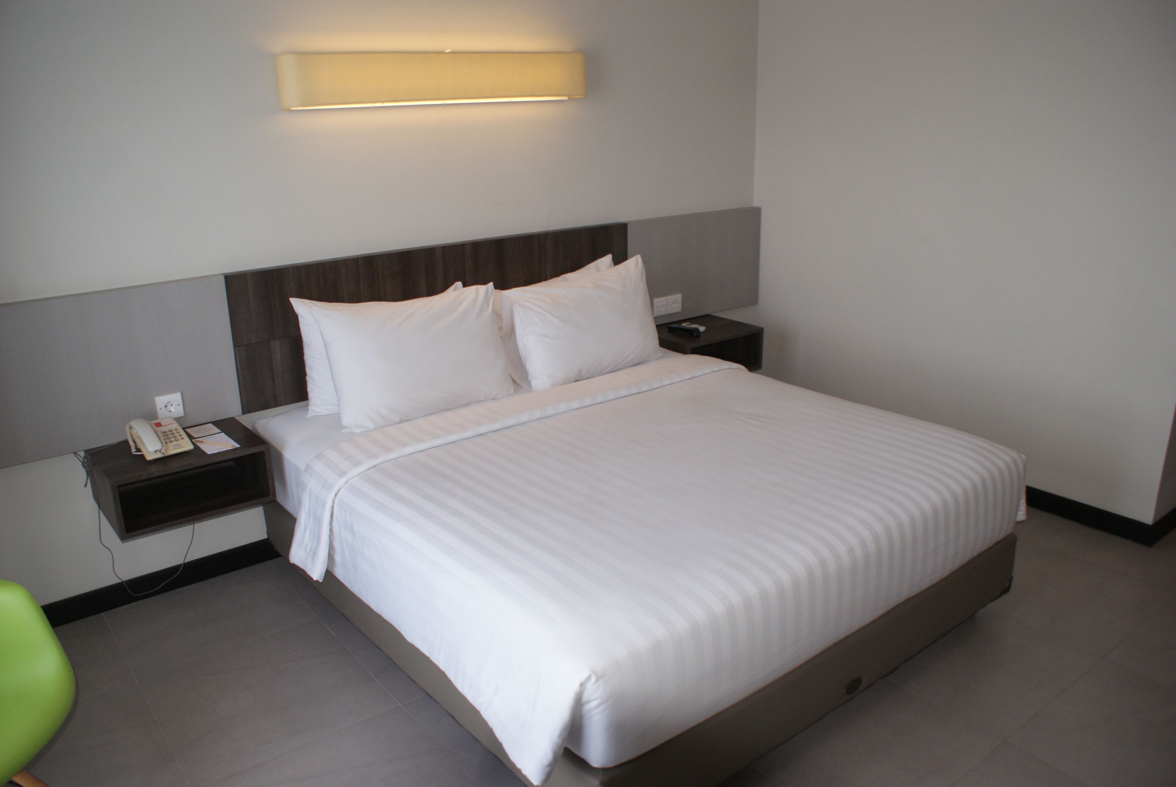 Una cama bien hecha con sábanas blancas, una mesita de noche con un teléfono y un bloc de notas, y una lámpara de pared sobre la cama.