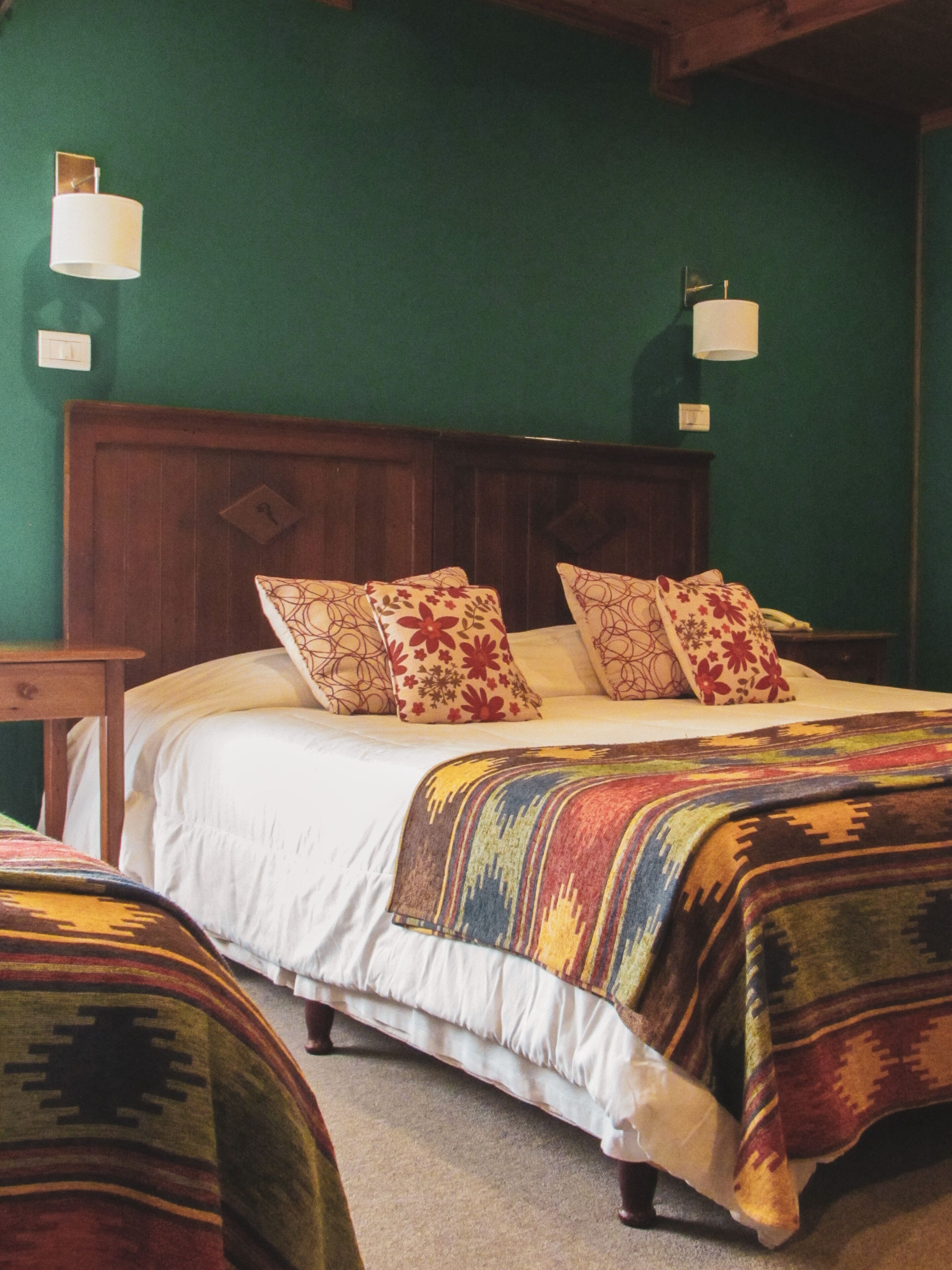 Una cama con cabecera de madera, ropa de cama blanca y almohadas estampadas. Hay dos mesitas de noche con lámparas. Las paredes son verdes y el suelo está cubierto de alfombra.