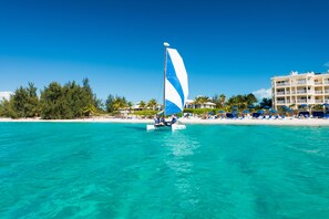 Un voilier sur une eau turquoise, avec une plage et des bâtiments d’hôtel en arrière-plan.