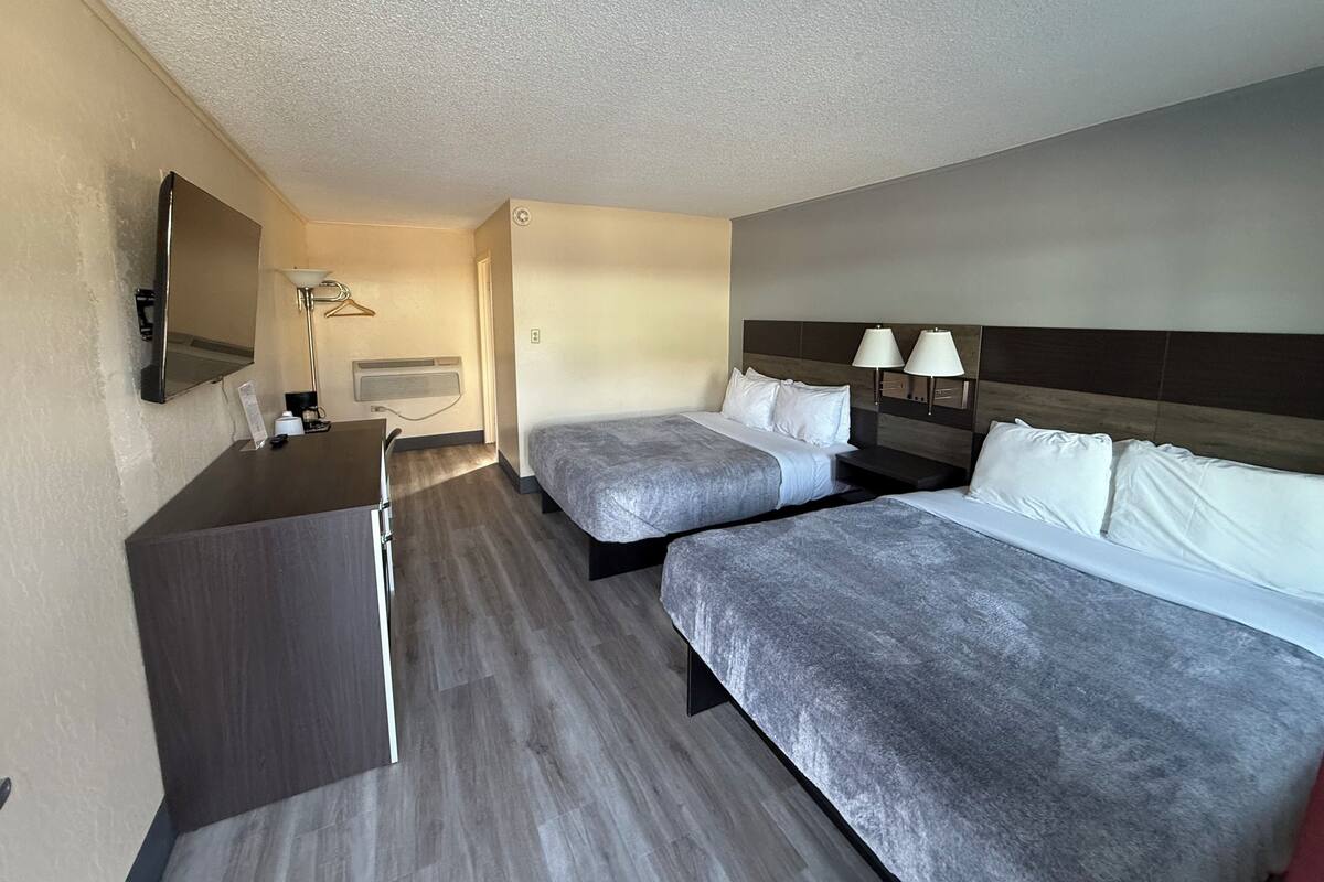 Quarto de hotel com duas camas, uma escrivaninha, um espelho e televisão na parede.