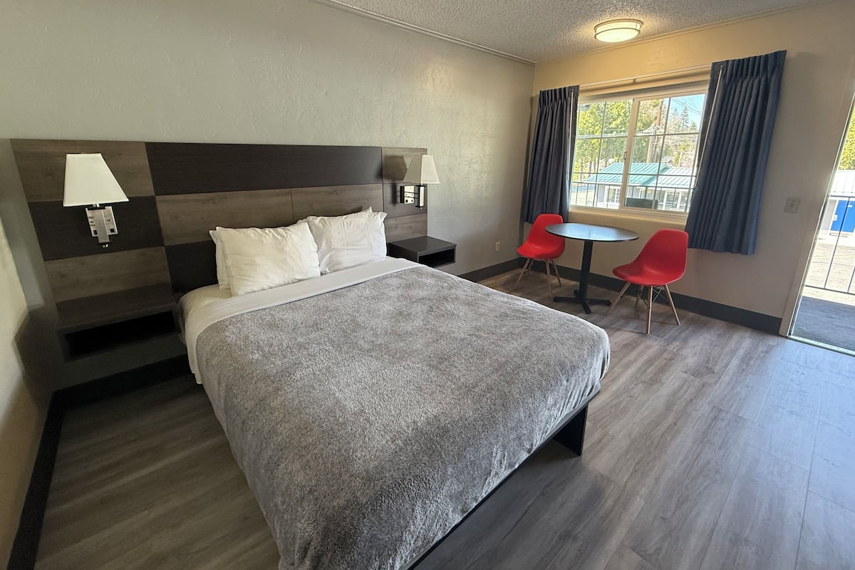 Quarto de hotel com uma cama grande, duas luminárias de cabeceira, uma mesa pequena com duas cadeiras vermelhas e uma janela com cortinas.