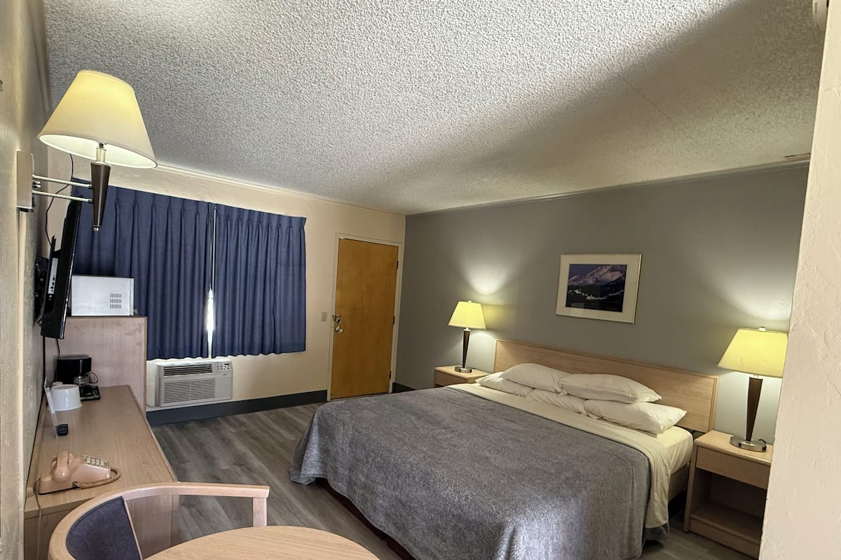 Quarto de hotel com cama, duas mesas de cabeceira com luminárias, escrivaninha com telefone, cadeira, TV fixada na parede e uma porta.