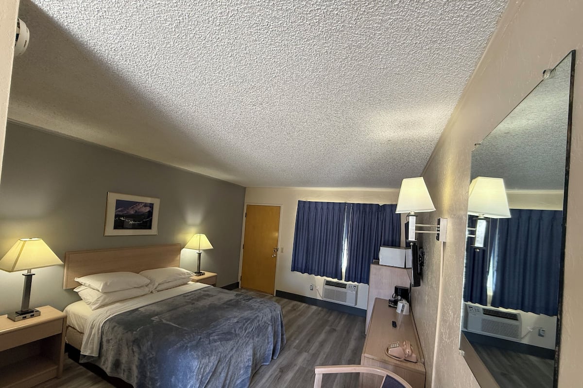 Quarto de hotel com uma cama, duas mesas de cabeceira com luminárias, uma escrivaninha com cadeira, um espelho e uma janela com cortinas.