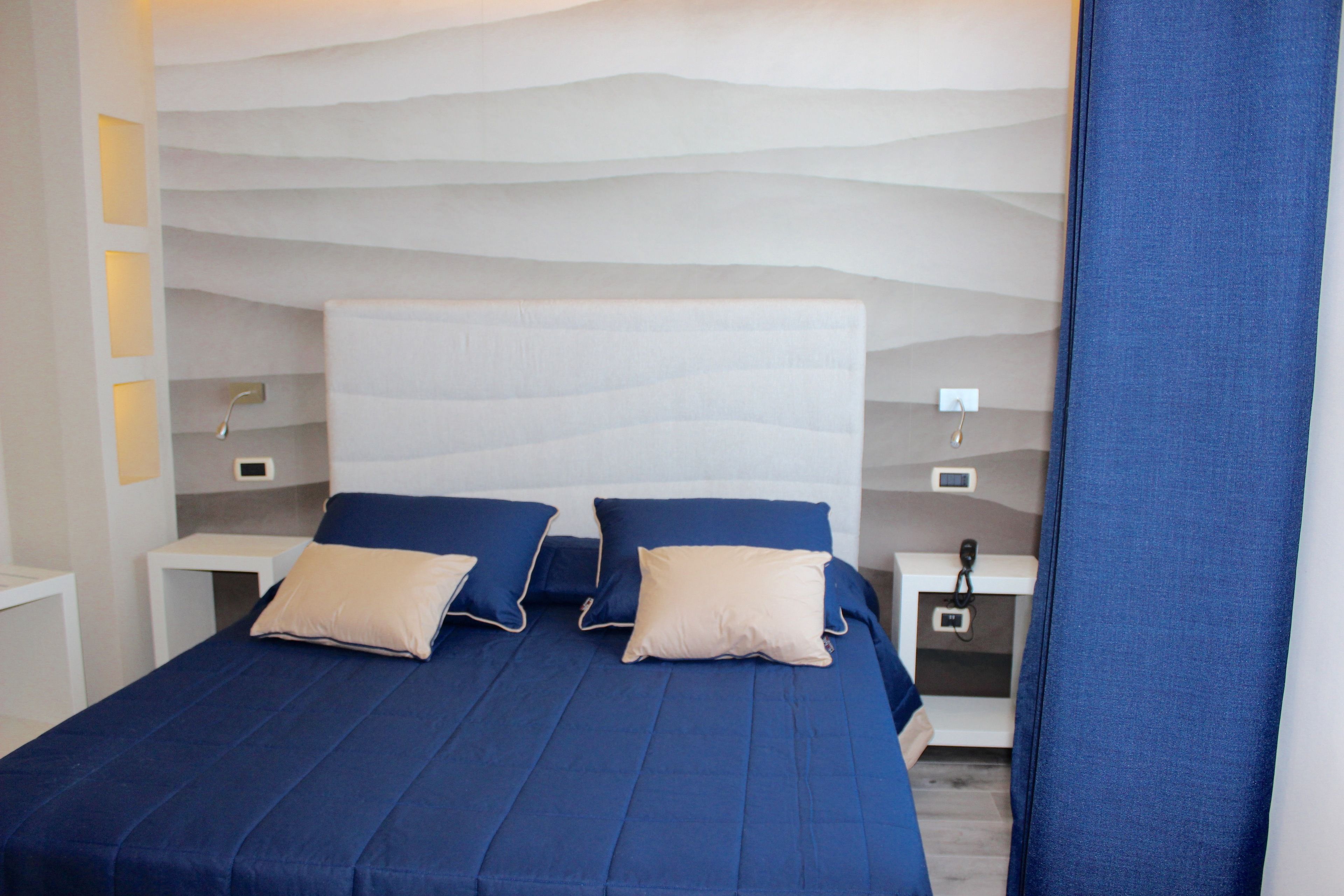A neatly made bed with blue bedding and two pillows, a bedside table with a lamp, and a wall-mounted control panel.
