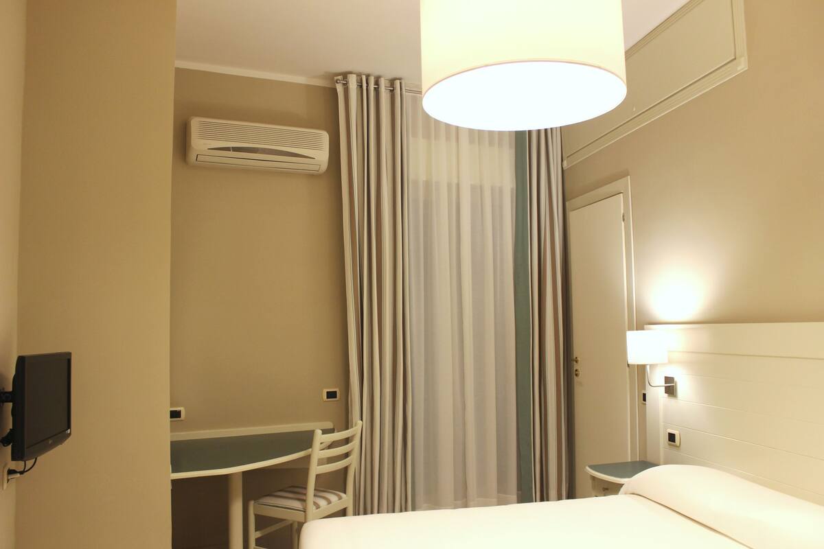 A hotel room with a bed, a desk, a chair, a curtain, an air conditioning unit, and a wall-mounted lamp.