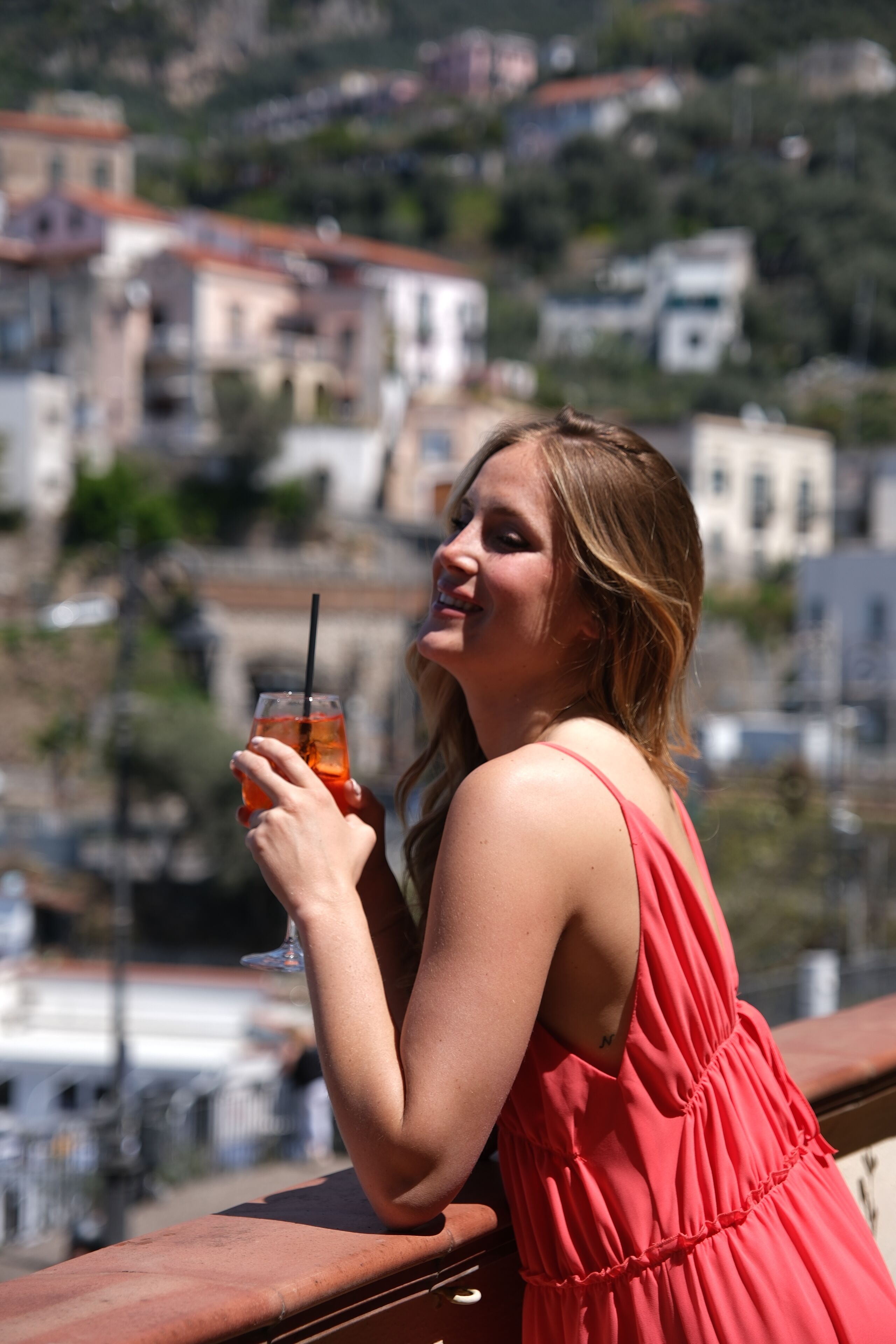 A person in a red dress holding a drink on a balcony with a city view in the background.