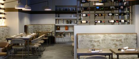 Un restaurante con varias mesas y sillas, una estantería con diferentes botellas de vino e iluminación colgante.
