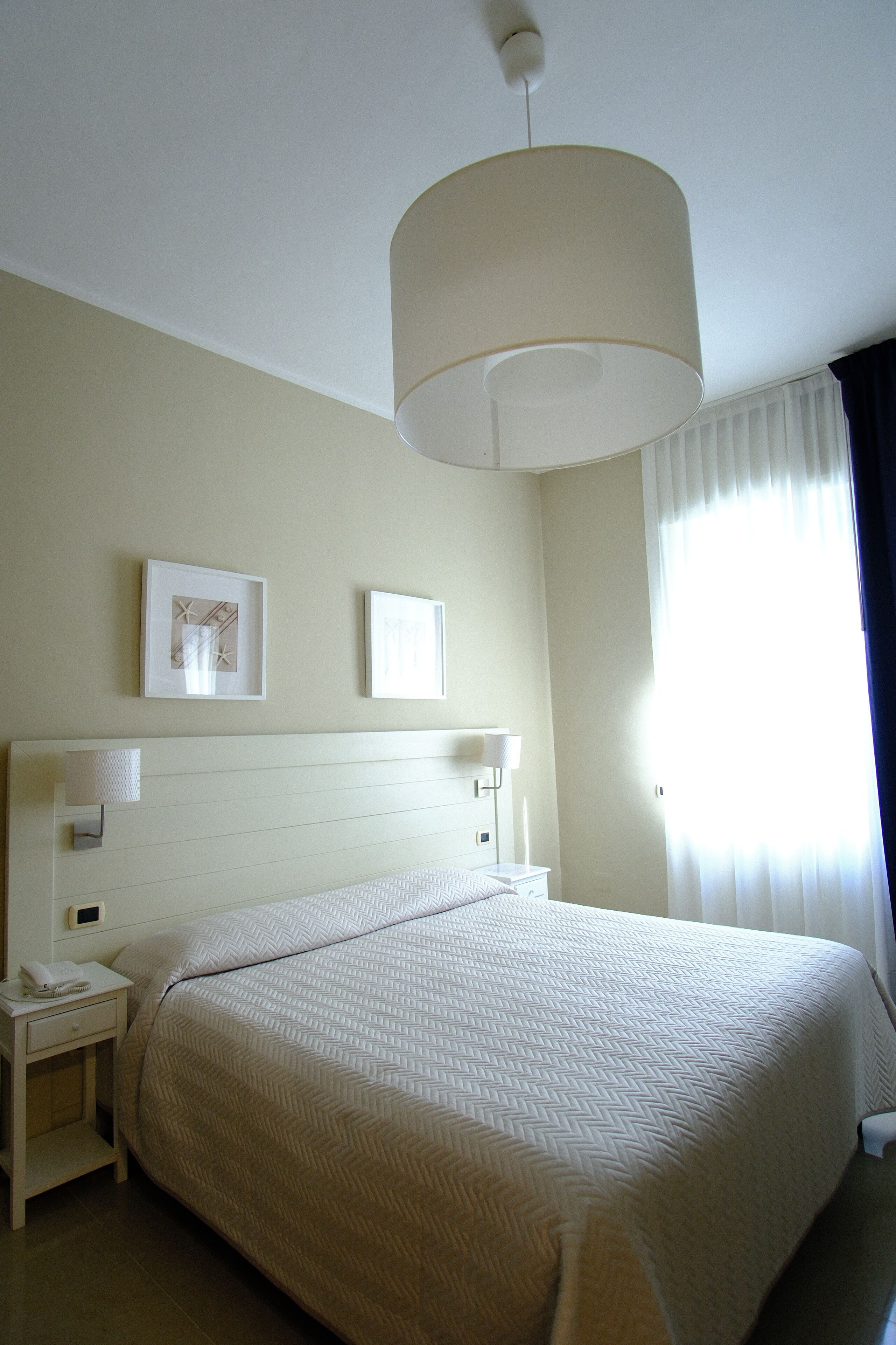 Una habitación de hotel con una cama grande, una mesita de noche con lámpara y dos cuadros en la pared.