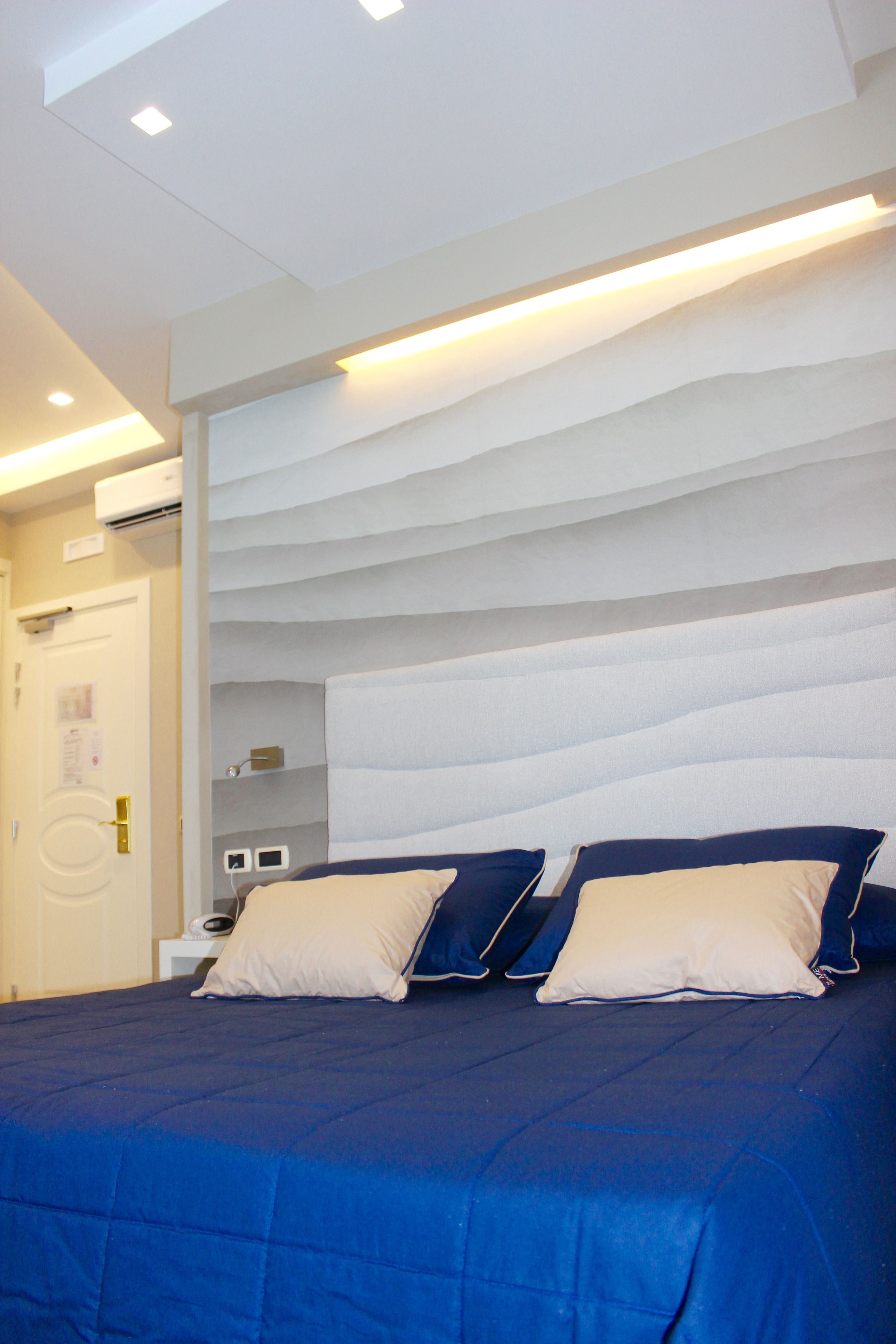 A hotel room with a bed, a white door, a white wall, and a ceiling with recessed lighting.