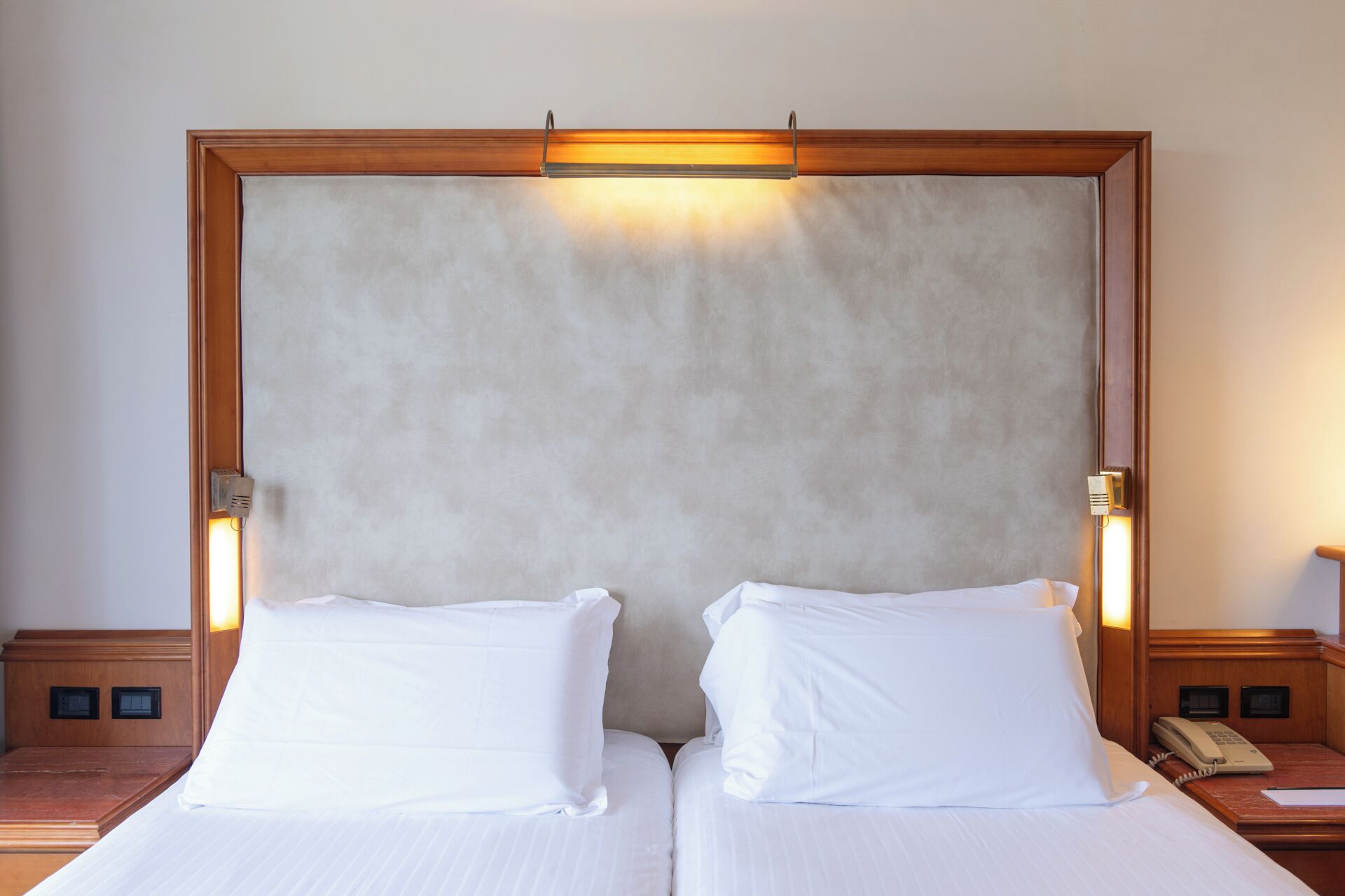 A hotel room with two single beds, a wooden headboard, a bedside telephone, and a light fixture above the headboard.