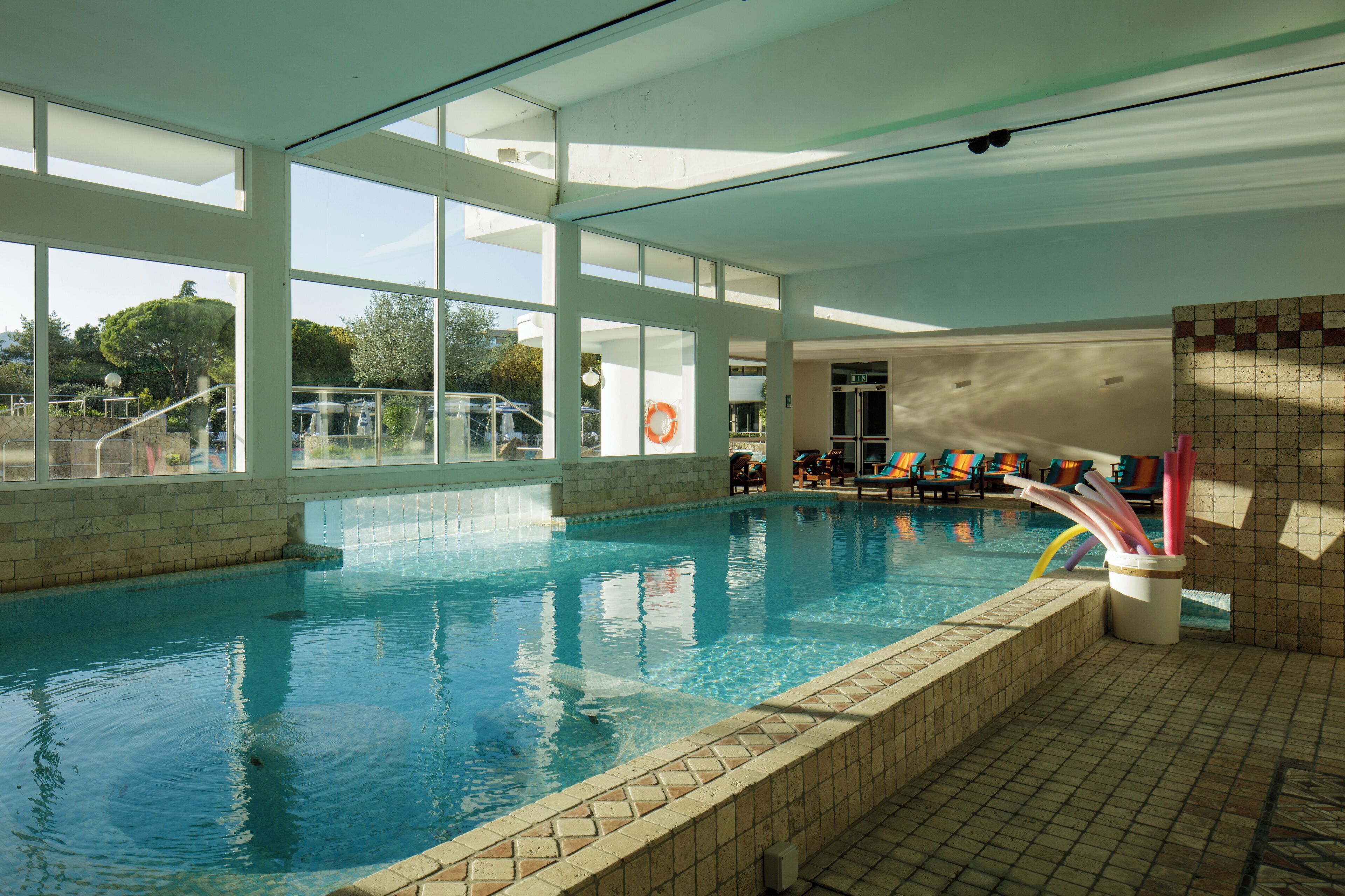 En indendørs swimmingpool med glasvægge, flisegulv og en redningskrans ved poolens kant.