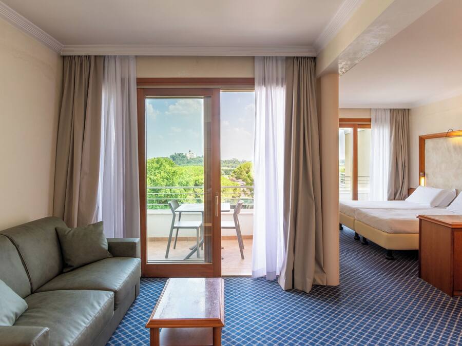 A hotel room with a sofa, a bed, a small table, and a balcony with a view of greenery.