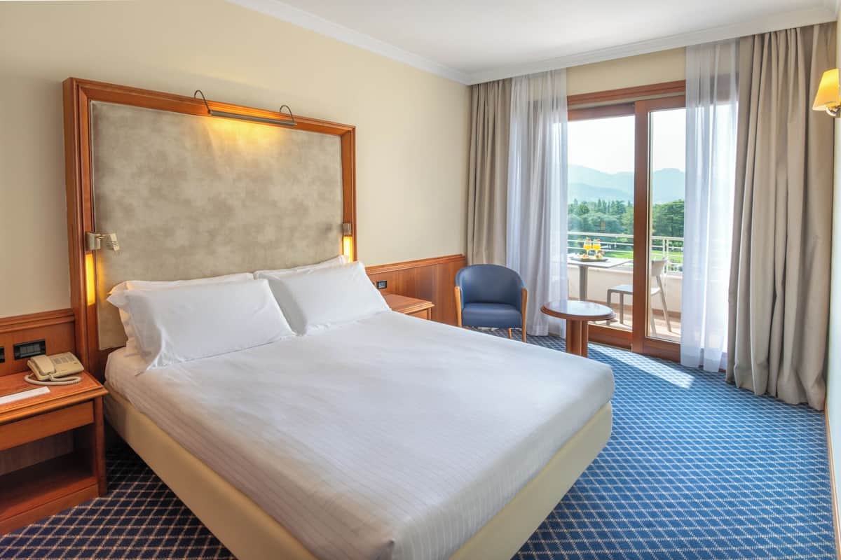 Une chambre d’hôtel avec un grand lit, un bureau avec un téléphone, une chaise et une petite table donnant sur l’extérieur.