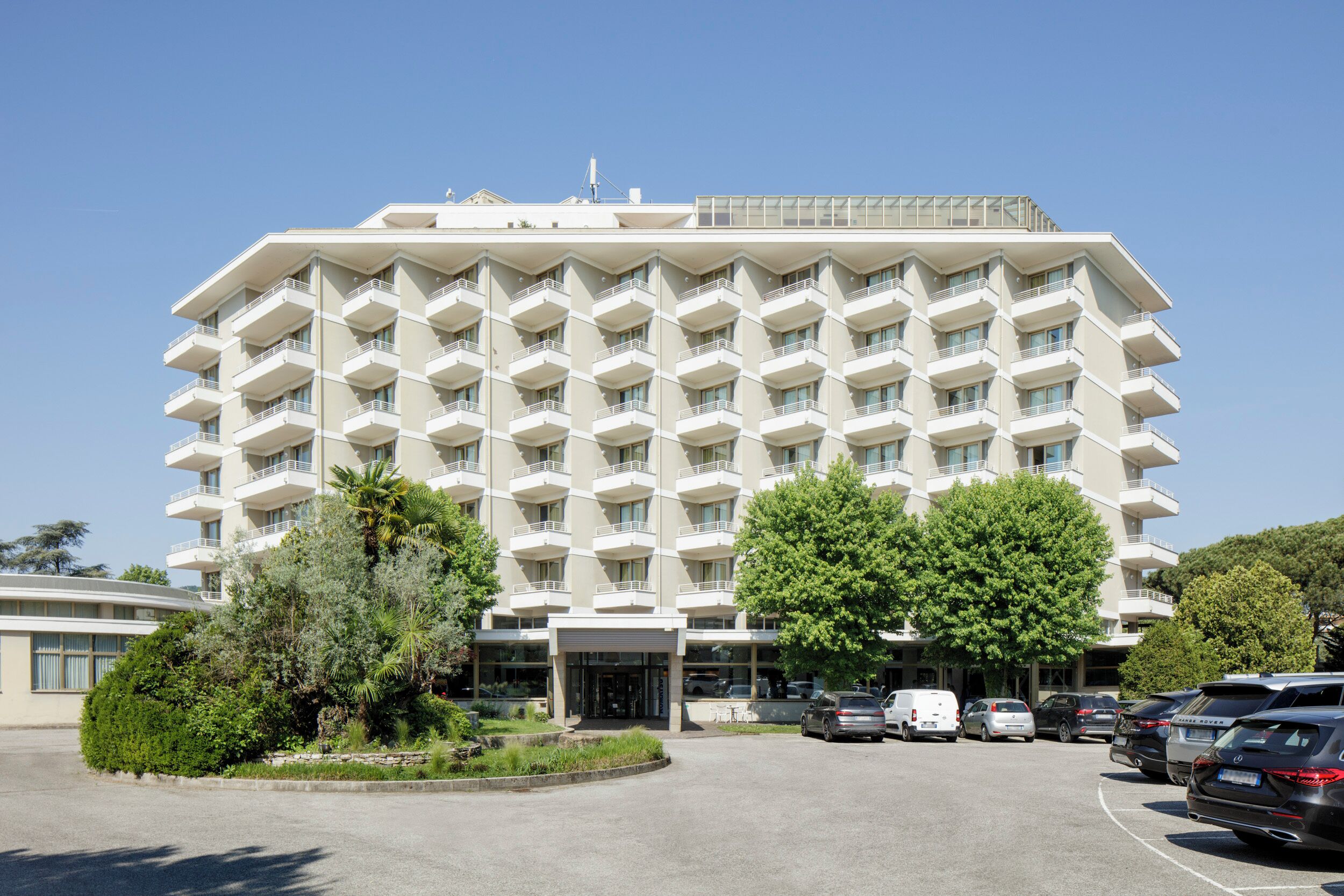 Un hotel a più piani con facciata simmetrica, un ingresso centrale e un parcheggio con diverse auto.