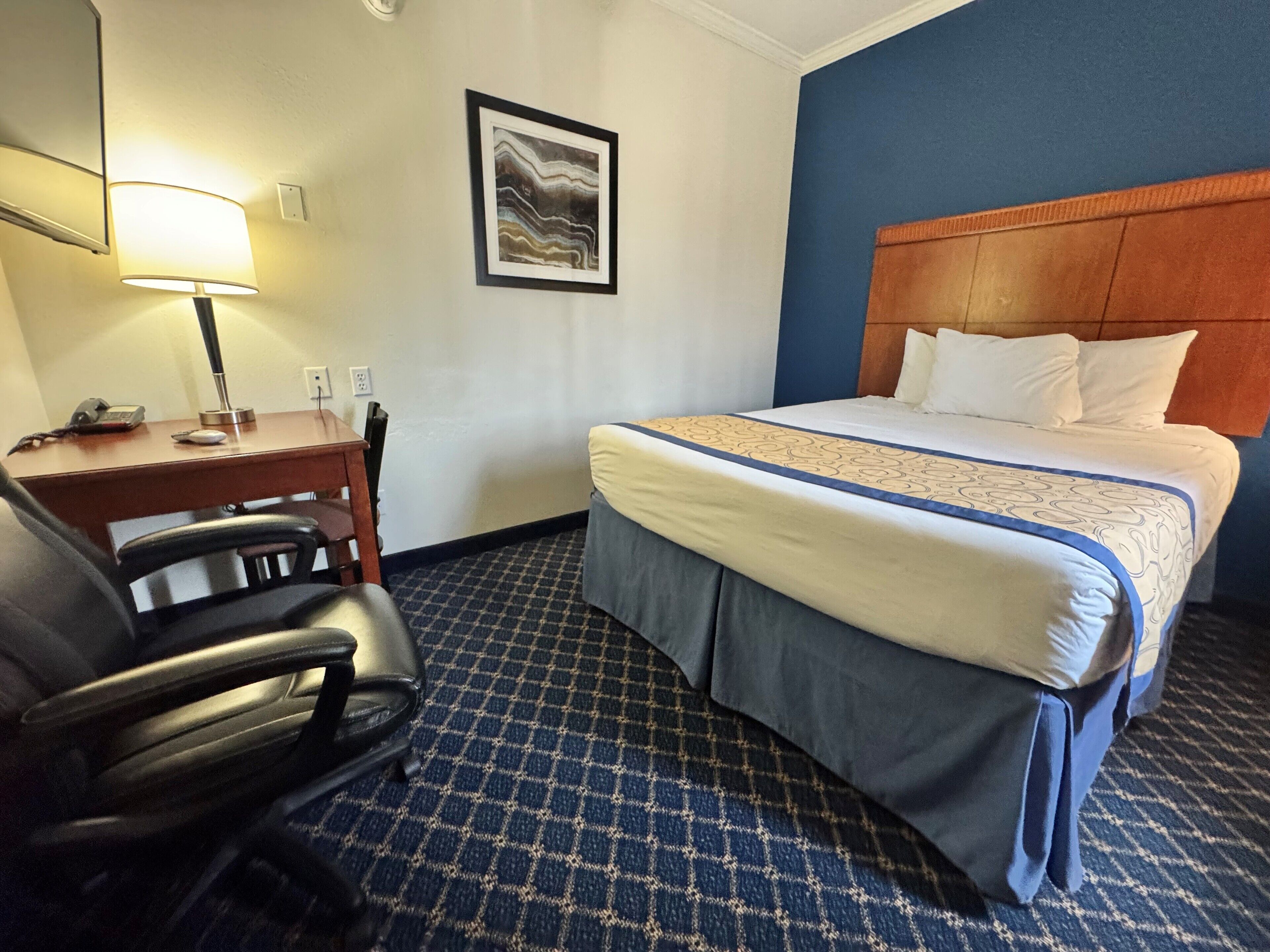 A hotel room with a bed, a desk with a lamp, a chair, and a framed picture on the wall.