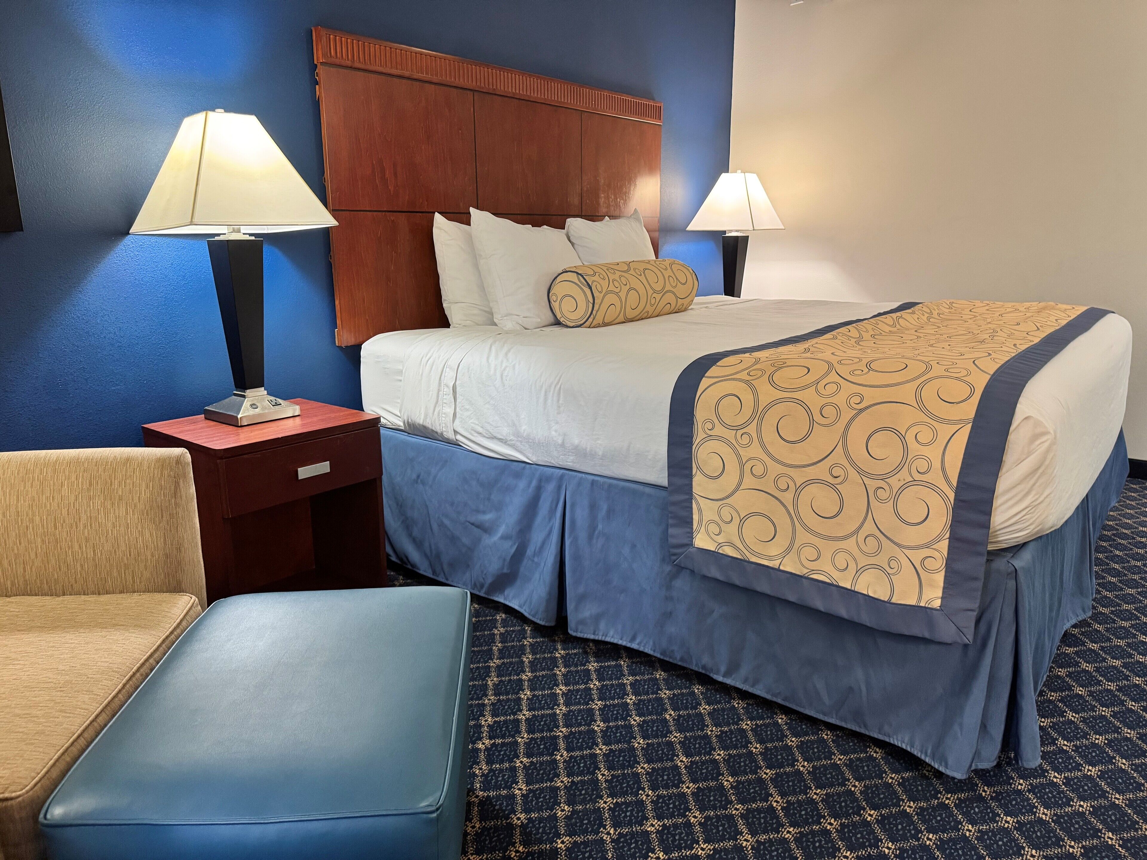 Habitación de hotel con una cama grande, dos lámparas de noche, un cabecero de madera, un sillón tapizado en azul y un otoman a juego.